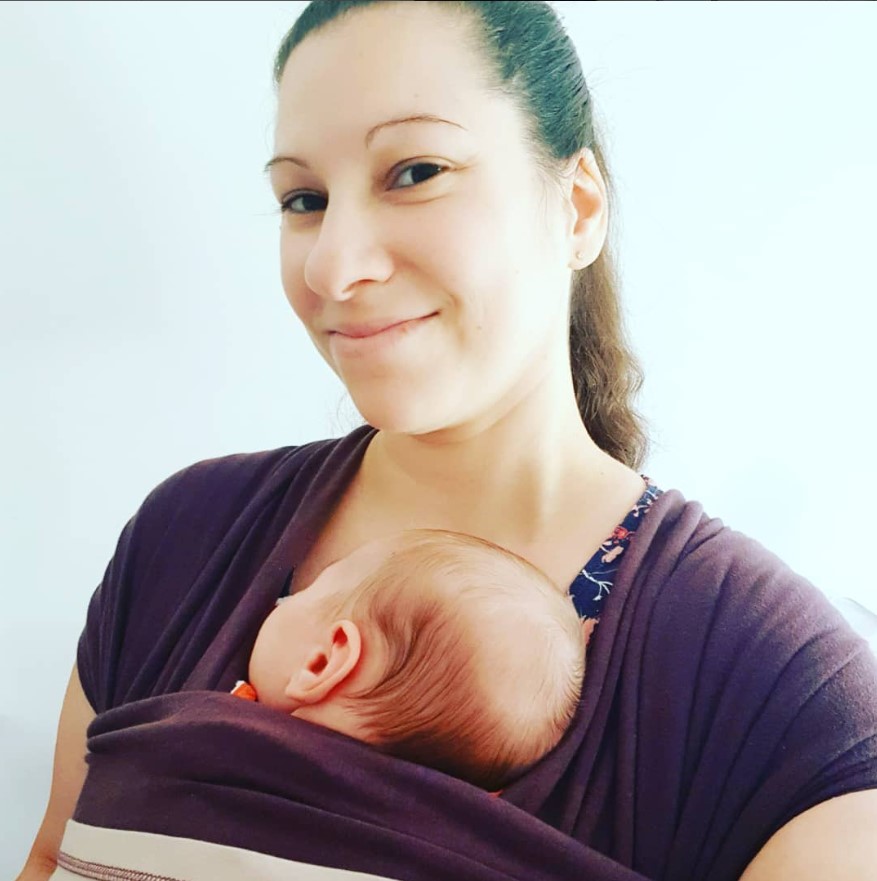Jeune maman portant son bébé endormi contre elle dans une écharpe de portage.
